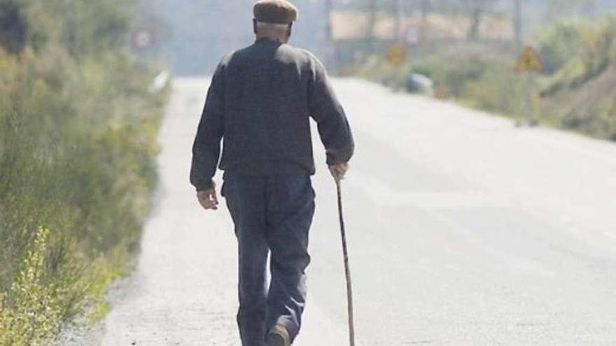 Un anciano camina por una carretera gallega apoyado en un bastón.