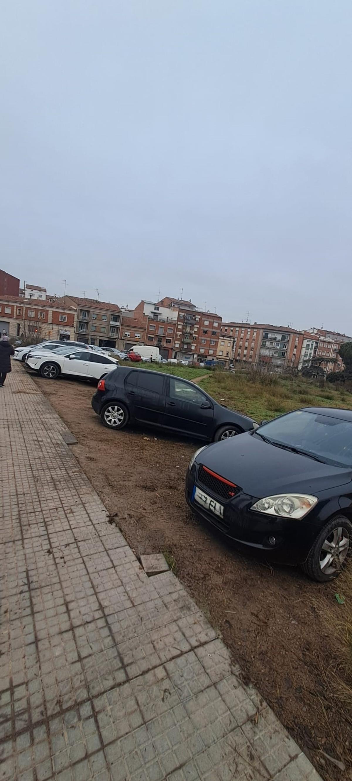 Cotxes aparcats al solar on es pot veure el tram de rajoles de la vorera que falta