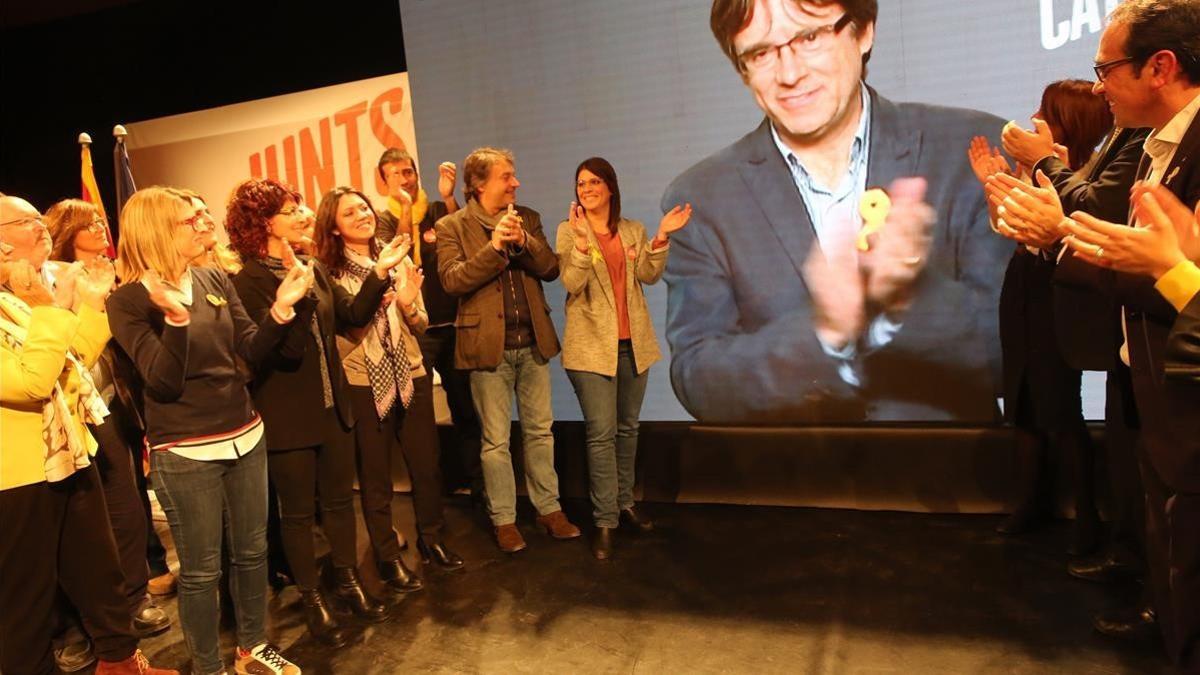 El escenario del mitin de JxCat en Girona, con Puigdemont en la pantalla y su esposa, aplaudiendo (en primera línea, la tercera por la izquierda).