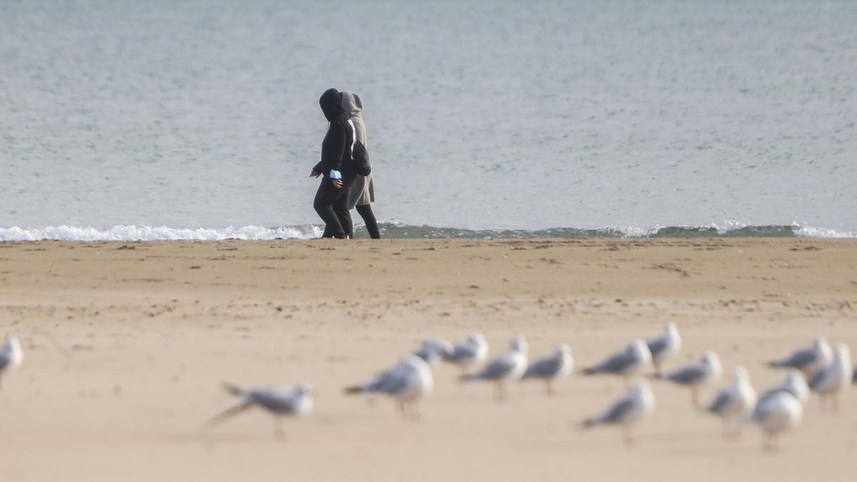 Dos personas pasean por la orilla de la playa abrigadas frente al frío en Valencia.