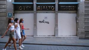 Jóvenes con mascarilla pasan por delante de la entrada de un hotel cerrado, en Barcelona.