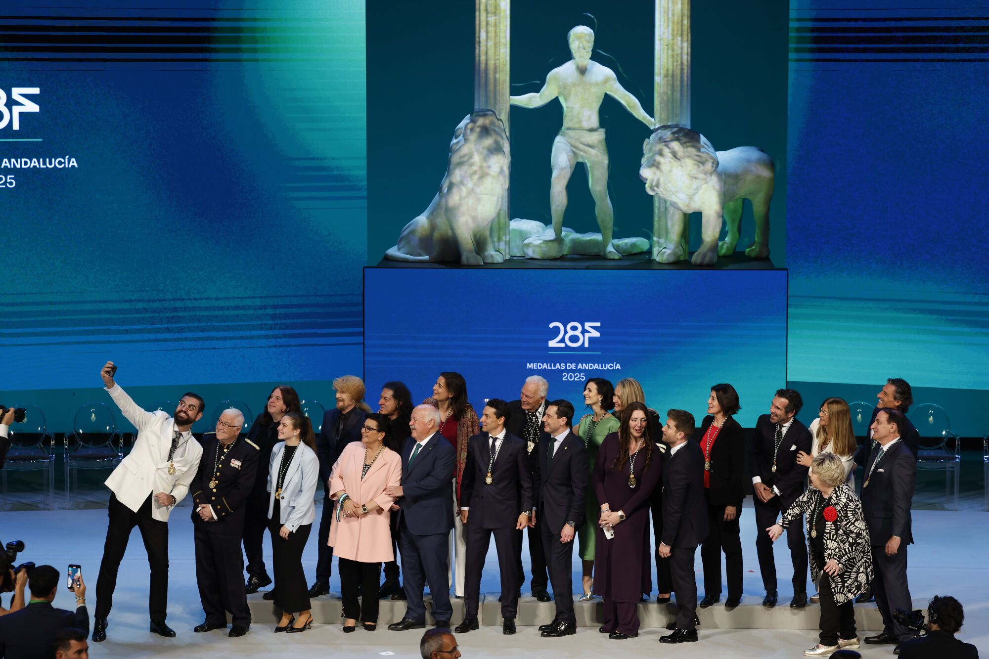 GRAFAND6621. SEVILLA, 28/02/2025.- El humorista y presentador Manu Sánchez, hace un selfie de todos los galardonados junto al presidente andaluz, Juanma Moreno (c), y el presidente de la Cámara autonómica, Jesús Aguirre (5i), durante el acto de entrega de las Medallas de Andalucía y las distinciones de Hijo/a Predilecto/a con motivo del Día de la comunidad andaluza , este viernes en Sevilla. EFE/Julio Muñoz