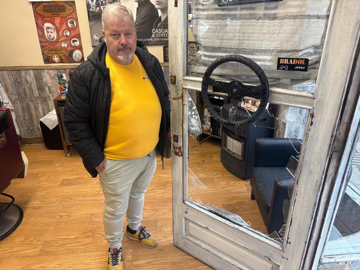 Rafael López junto a la puerta de su peluquería de Sama con el cristal roto por los ladrones