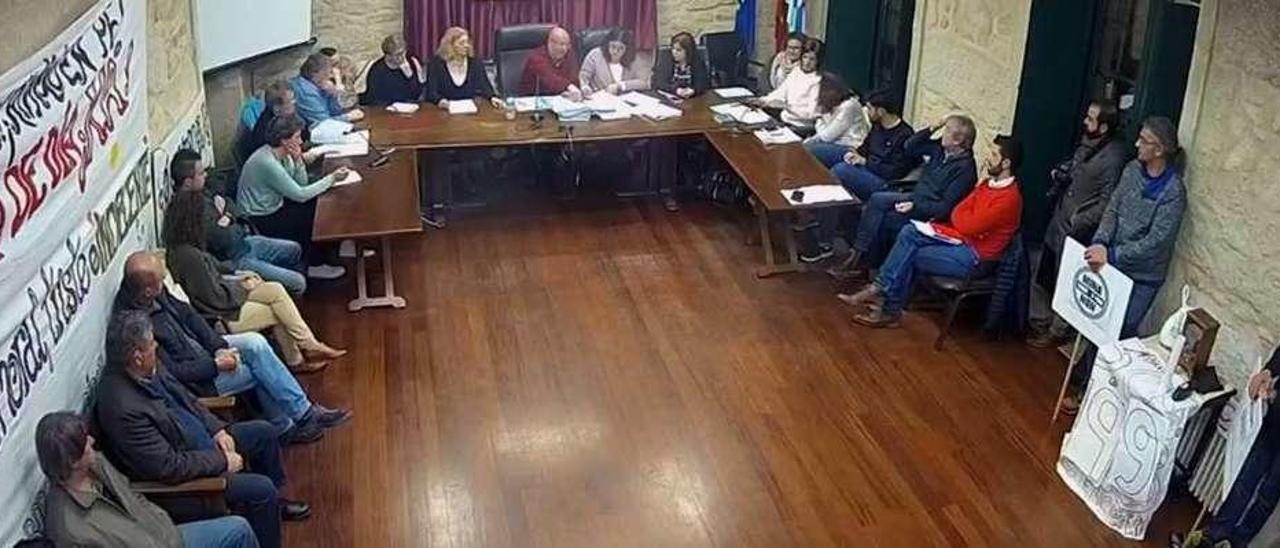 Vecinos encerrados por un centro de día y emisión en directo  |  Esta foto corresponde a una captura de pantalla de la proyección en directo -vía YouTube- del pleno de la Corporación municipal de Rianxo. De este modo el municipio arousano se estrena en este tipo de proyecciones, con el propósito de ofrecer mayor transparencia y facilitar a sus vecinos el acceso de primera mano a la información y el día a día de la actividad municipal. Al mismo tiempo es posible comprobar -como se aprecia en las pancartas colocadas en los laterales del salón de plenos-, que sigue la lucha de los vecinos rianxeiros para conseguir un centro de día. Es una pelea iniciada hace ya unos años que llevó a los ciudadanos a organizarse para permanecer encerrados en el consistorio y reivindicar así dicha infraestructura, además de ejercer otras medidas de presión.