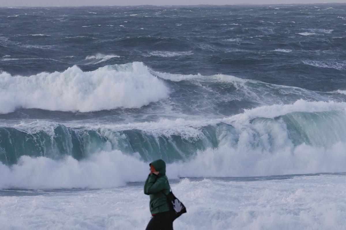 Una persona camina delante del fuerte oleaje esta tarde en la costa de Muxía, en la gallega Costa de la Muerte. Tras el paso ayer lunes de la borrasca Gérard, la parte más adversa de este temporal invernal corresponderá a la borrasca Fien, que a partir de este martes mantiene activado el aviso rojo en Galicia por olas de 9 metros y en otras 12 comunidades en naranja por nieve en cotas bajas, lluvias y fuerte viento.