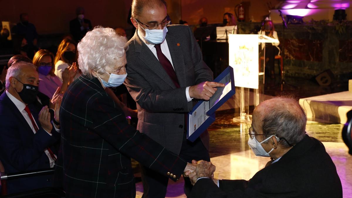 El presidente aragonés, Javier Lambán, entrega el reconocimiento a Ana María Cortés, que estrecha la mano del presidente Hipólito Gómez de las Roces, este 8 de marzo en la Sala de la Corona del Pignatelli.