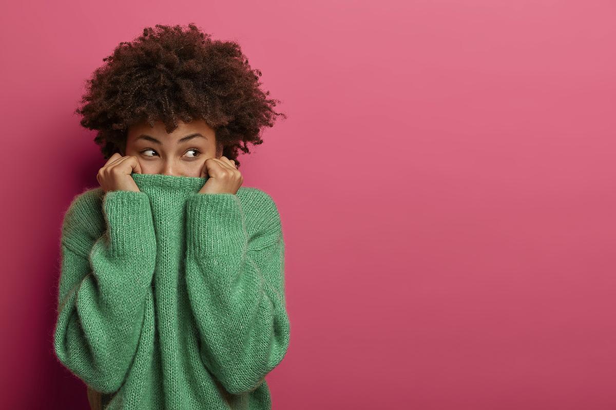 Positive curly young woman pulls green sweater over head, has fun and hides face, looks from underneath, disappears in her clothes, gazes gladfully, isolated on pink background, copy space area