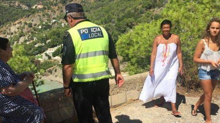 El agente de la Policía Local de Sant Josep indica a dos turistas que no pueden seguir por el camino, mientras Maria descansa a la sombra de un pino.