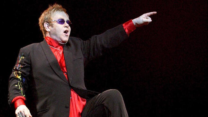 Elton John, subido a una silla y su piano, señala al público durante un concierto.