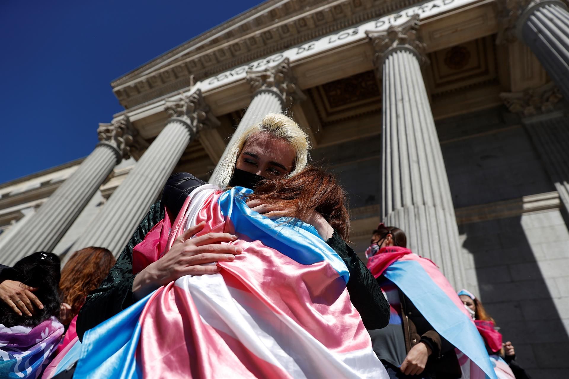 Dos personas envueltas en la bandera trans se abrazan a las puertas del Congreso de los Diputados.