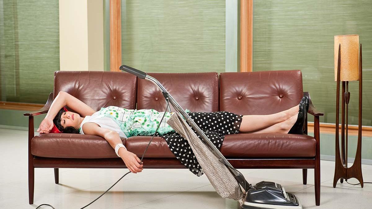 Mujer ama de casa con delantal tumbada agotada con aspiradora sobre un sofá
