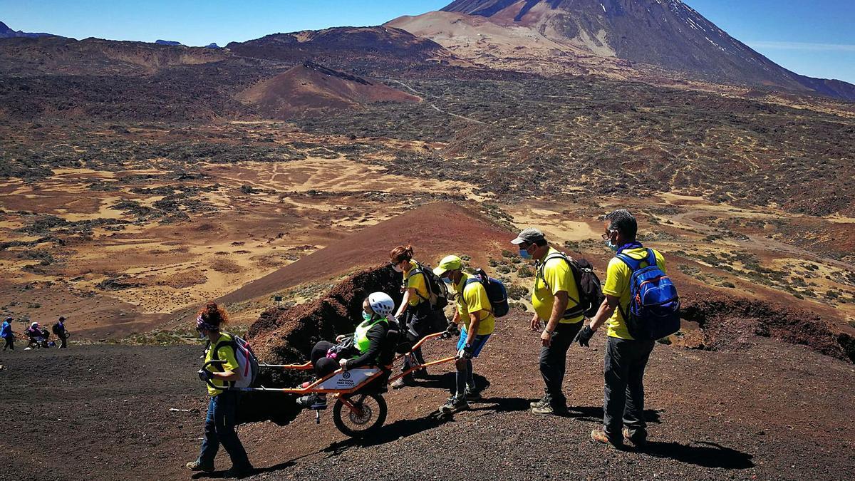 Voluntarios de Montaña para todos llevan a una persana con discapacidad sobre una Joëlette en una ruta de senderismo con el Teide al fondo.