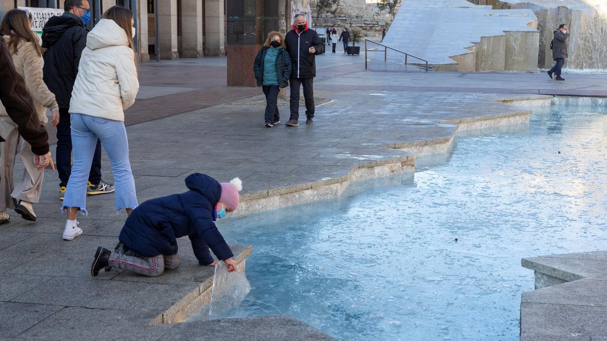 ZARAGOZA, 23/01/2022.- Una niña coge un trozo de hielo de la fuente de La Hispanidad, helada por las bajas temperaturas de esta noche pasada. La Agencia Estatal de Meteorología (Aemet) anuncia para este domingo en Aragón, heladas generalizadas, más intensas en fondos de valles del Pirineo y de la Ibérica, donde podrán ser localmente fuertes. EFE/JAVIER BELVER