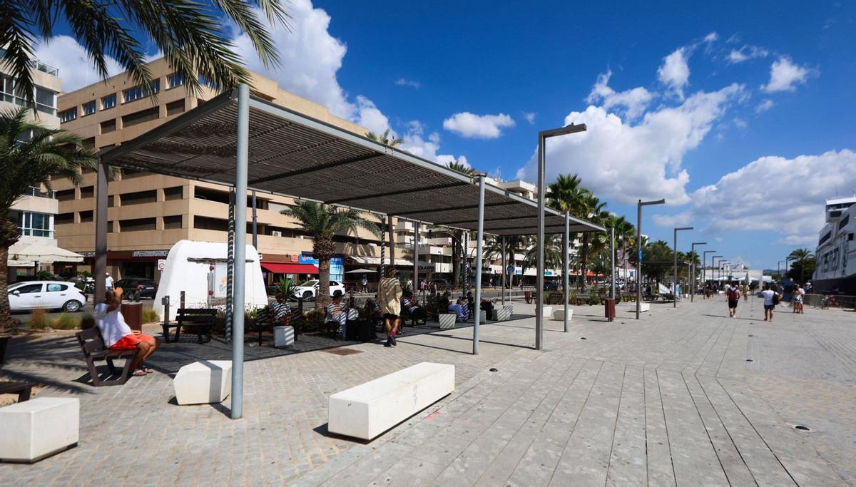 Varias personas sentadas, a la izquierda, en bancos con sombra frente a las barcas de Formentera. | TONI ESCOBAR