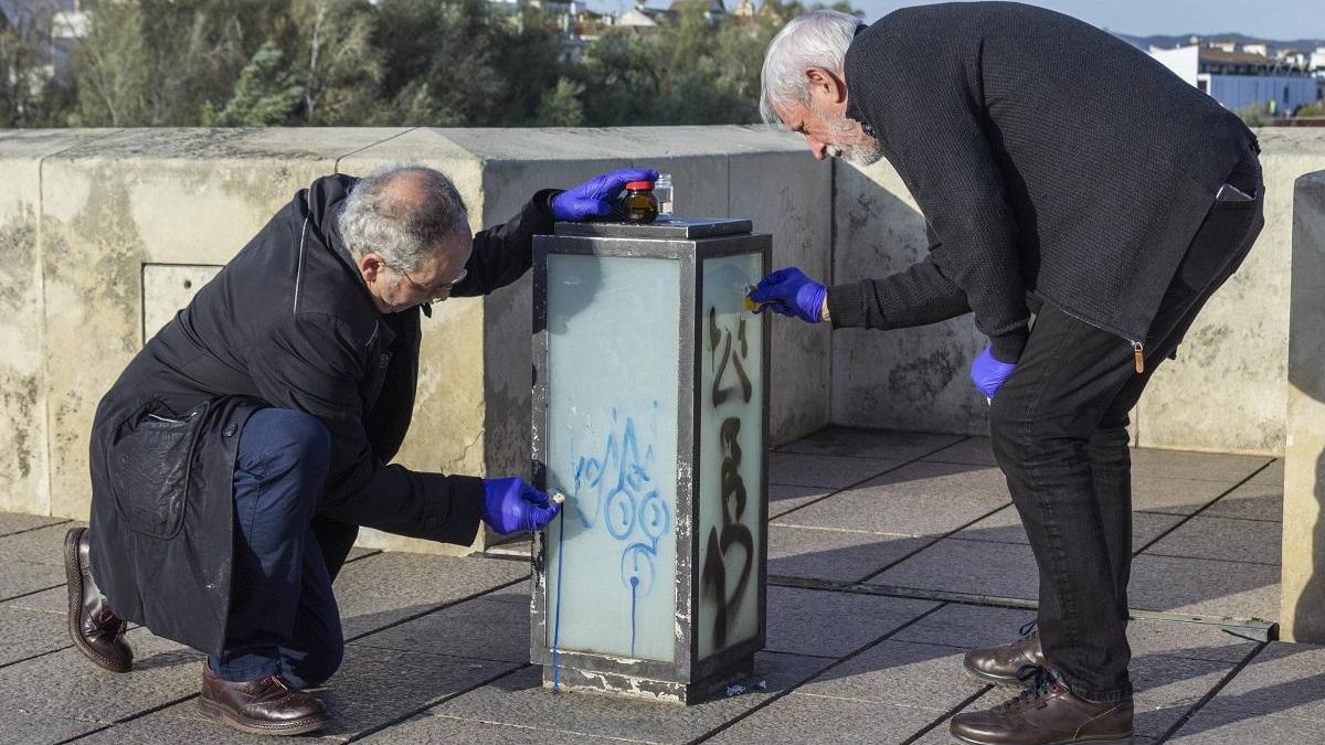 Dos vecinos limpian de grafitis una de las farolas del Puente Romano.