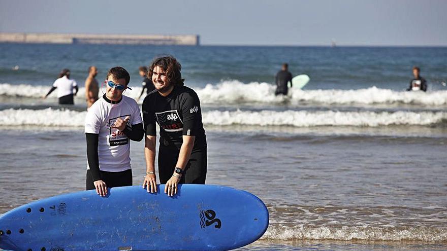 Por la izquierda, Sam Daly junto a su monitor, Daniel Peláez. De fondo, la clase de surf en el arenal gijonés. | Ángel González