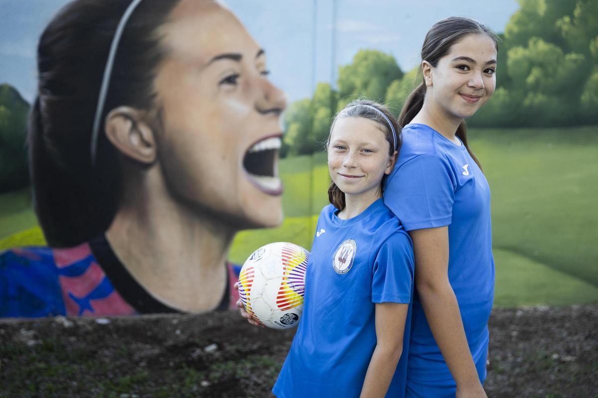 Gina Cruells y Laura Solorzano delante del mural del campo de fútbol Aitana Bonmatí