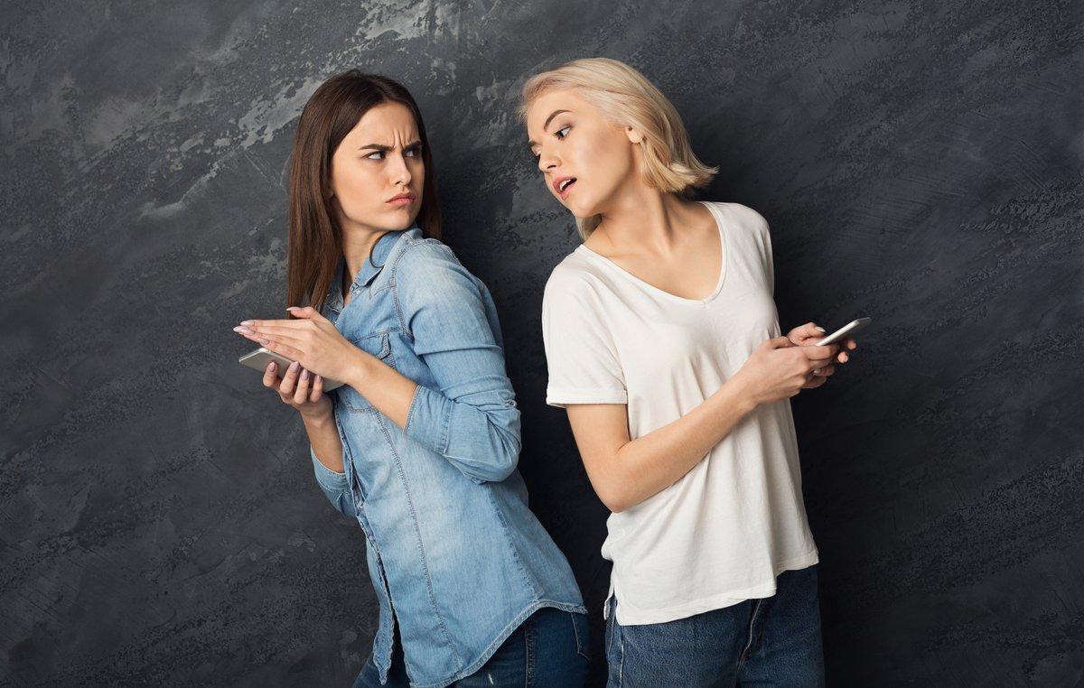 IMAGEN BAJADA DE 123RF EL 4 DE MARZO DEL 2019. CHICAS ESPIANDO LOS MOVILES DE LA OTRA. Girls prying into each other phones at gray background. Spying concept. ESPIAR, CELOS, ENVIDIA, MIRAR, CHAFARDEAR, CHAFARDEO, MÃVIL, CHICAS. FOTO DE KONSTANTIN POSTUMITENKO. 123RF.