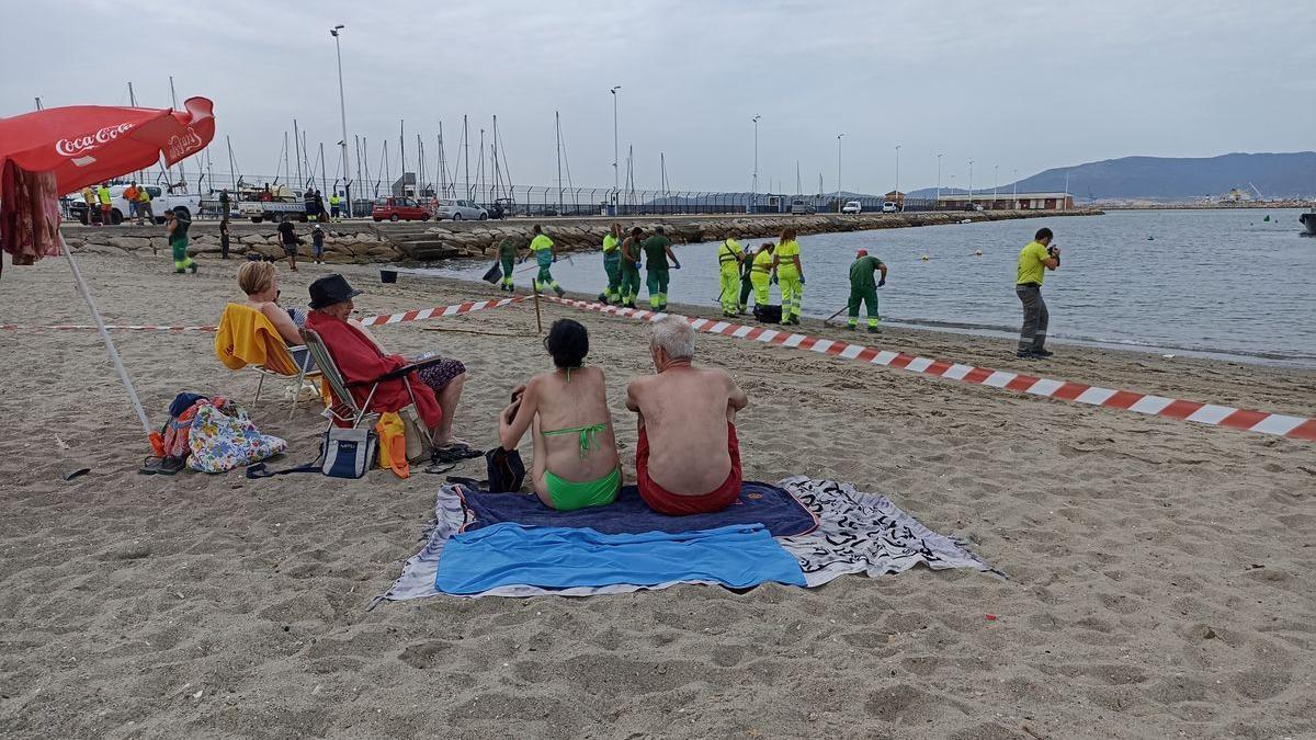 Turistas observan como operarios de La Línea retiran restos de aceite de la Playa de Poniente.
