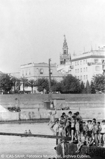 Jóvenes participan en la cucaña con la Giralda al fondo y el desaparecido edificio de la clínica del doctor Zbikowski (Década de 1960)