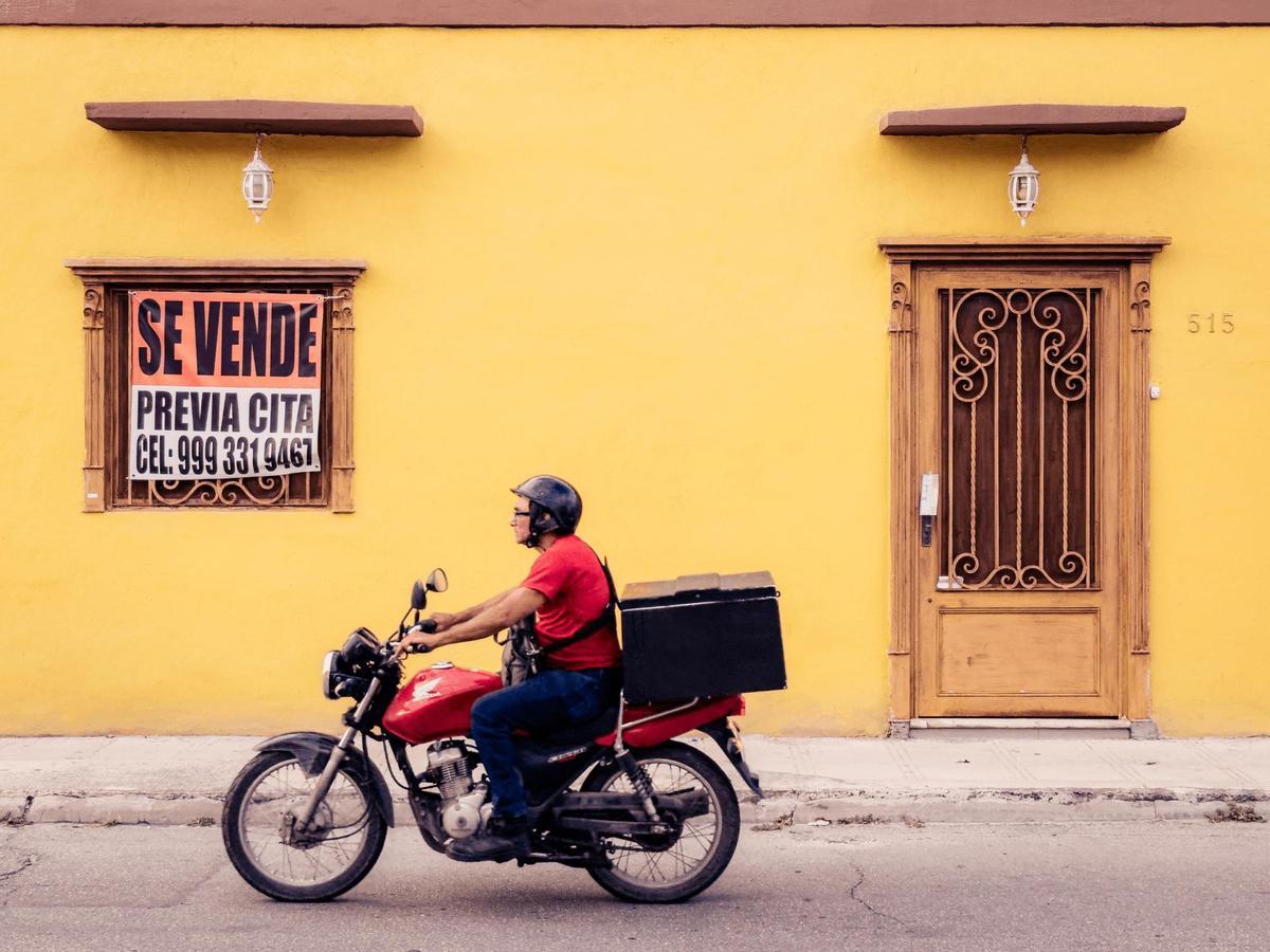 Mérida, México. Un repartidor en moto cruza frente a una fachada amarilla, una de las tantas casas de colores que le dan identidad a las calles de la Ciudad Blanca, apodo que Mérida recibe por su arquitectura colonial y su tradición de edificios impecablemente mantenidos. (iPhone 14 Pro)