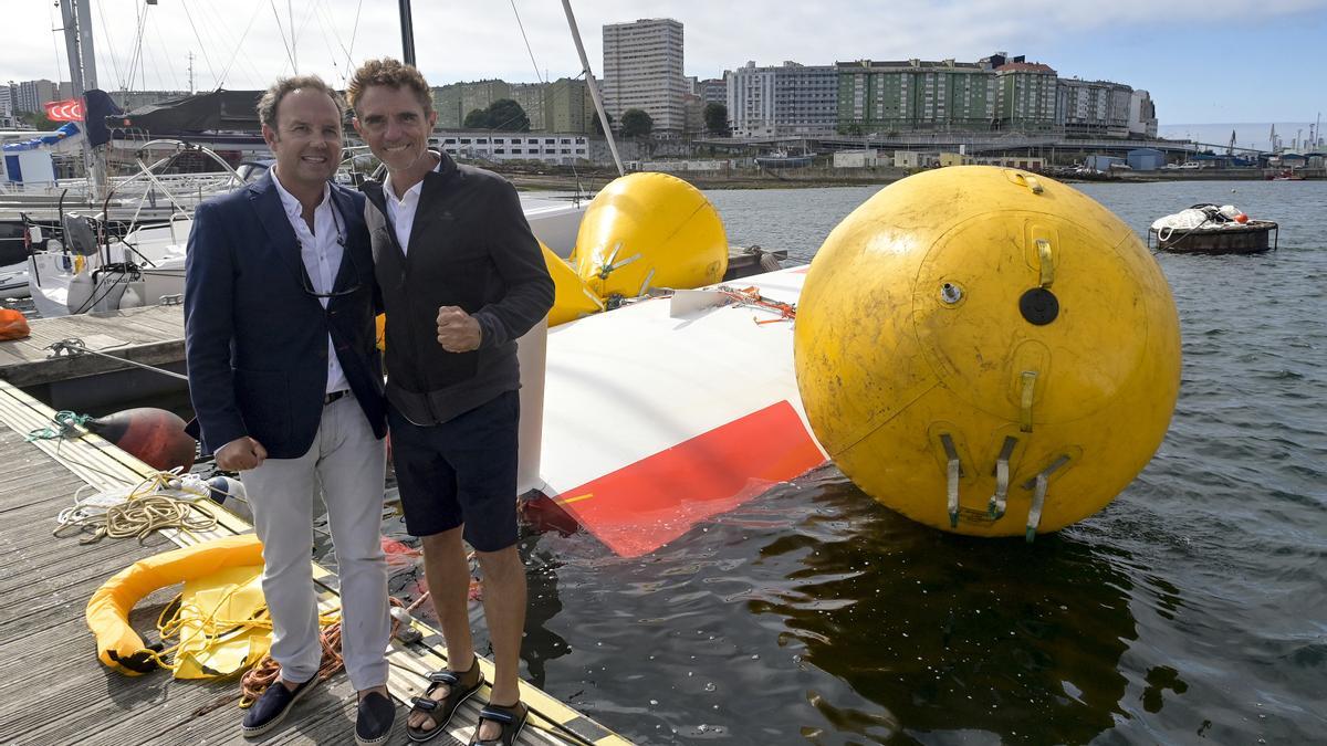 El navegante francés Laurent Camprubi (d), acompañado por el Jefe de Salvamento Marítimo Finisterre, Manuel Capeáns (i), posan delante del velero volcado y amarrado en el pantalán del puerto marítimo de Oza.