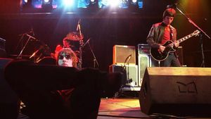 Liam Gallagher sentado en el escenario mientras Noel toca la guitarra.