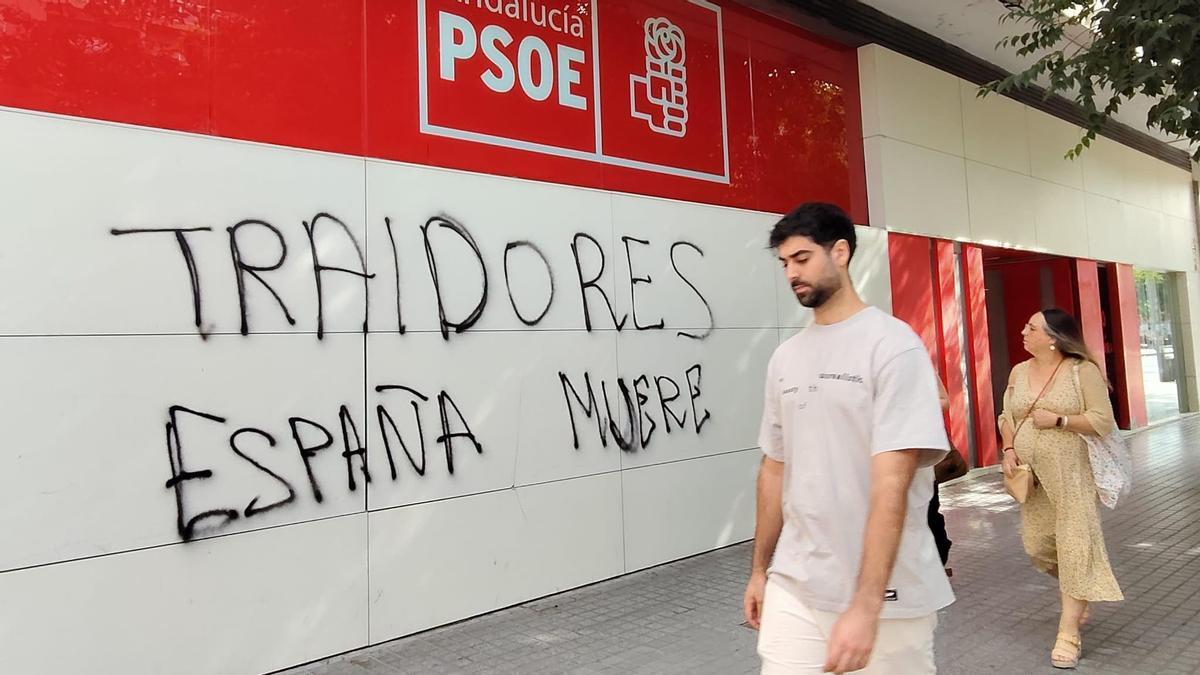 Dos personas caminan junto a la pintada en la sede del PSOE de Córdoba, en avenida del Aeropuerto.