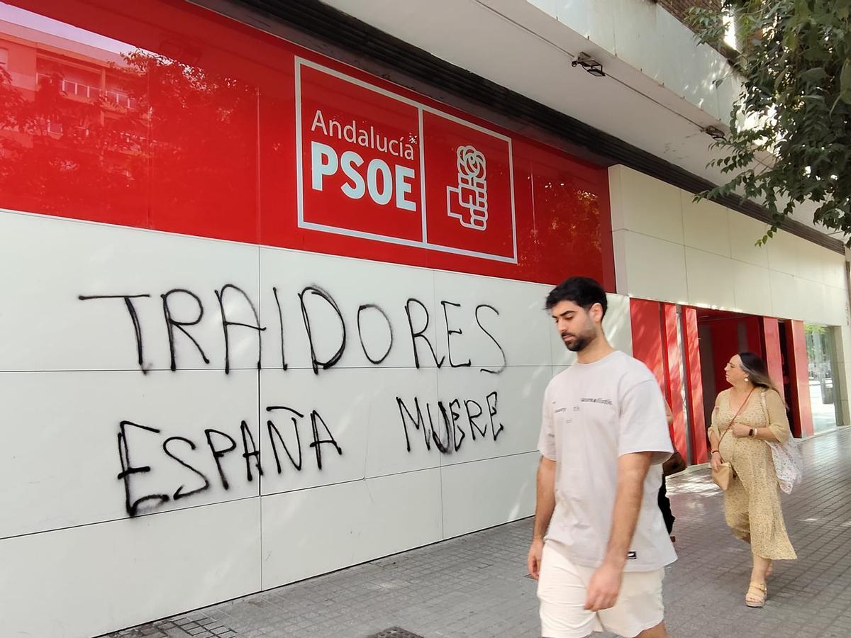 Dos personas caminan junto a la pintada en la sede del PSOE de Córdoba, en avenida del Aeropuerto.