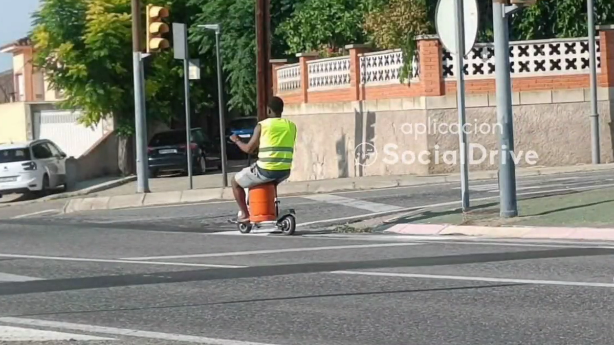 Un hombre circula en patinete eléctrico sentado sobre una bombona de butano