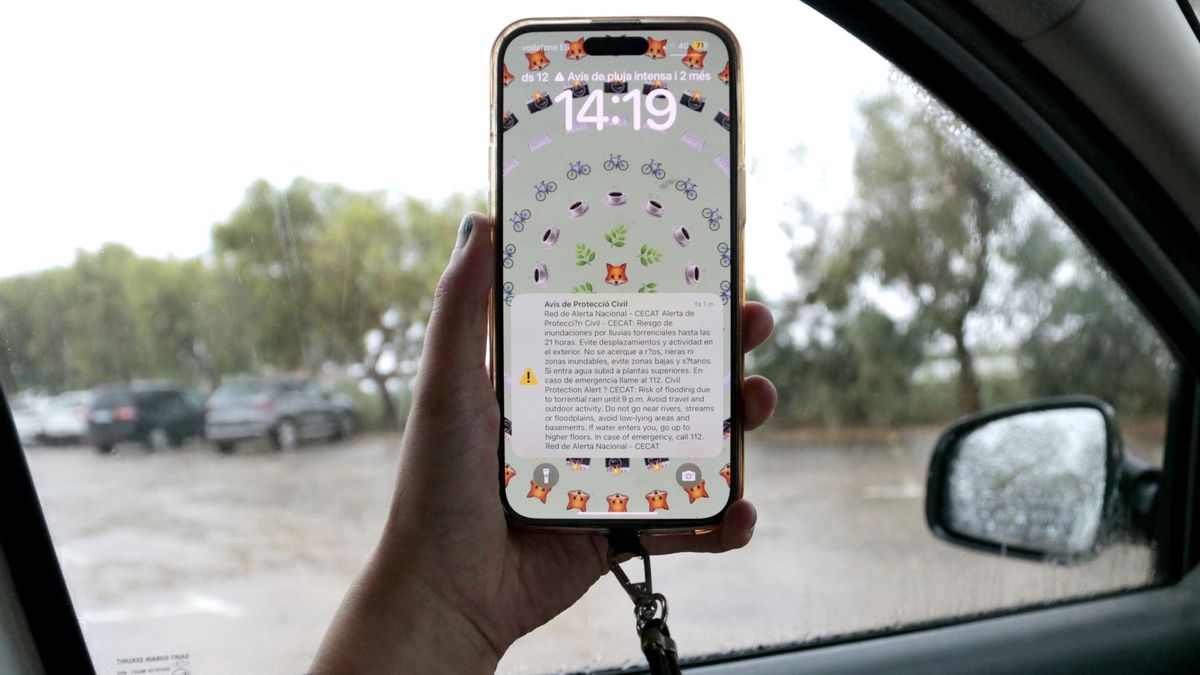 Un telèfon mòbil, amb l'alerta de Protecció Civil, i un carrer ple d'aigua de pluja al fons, a Amposta