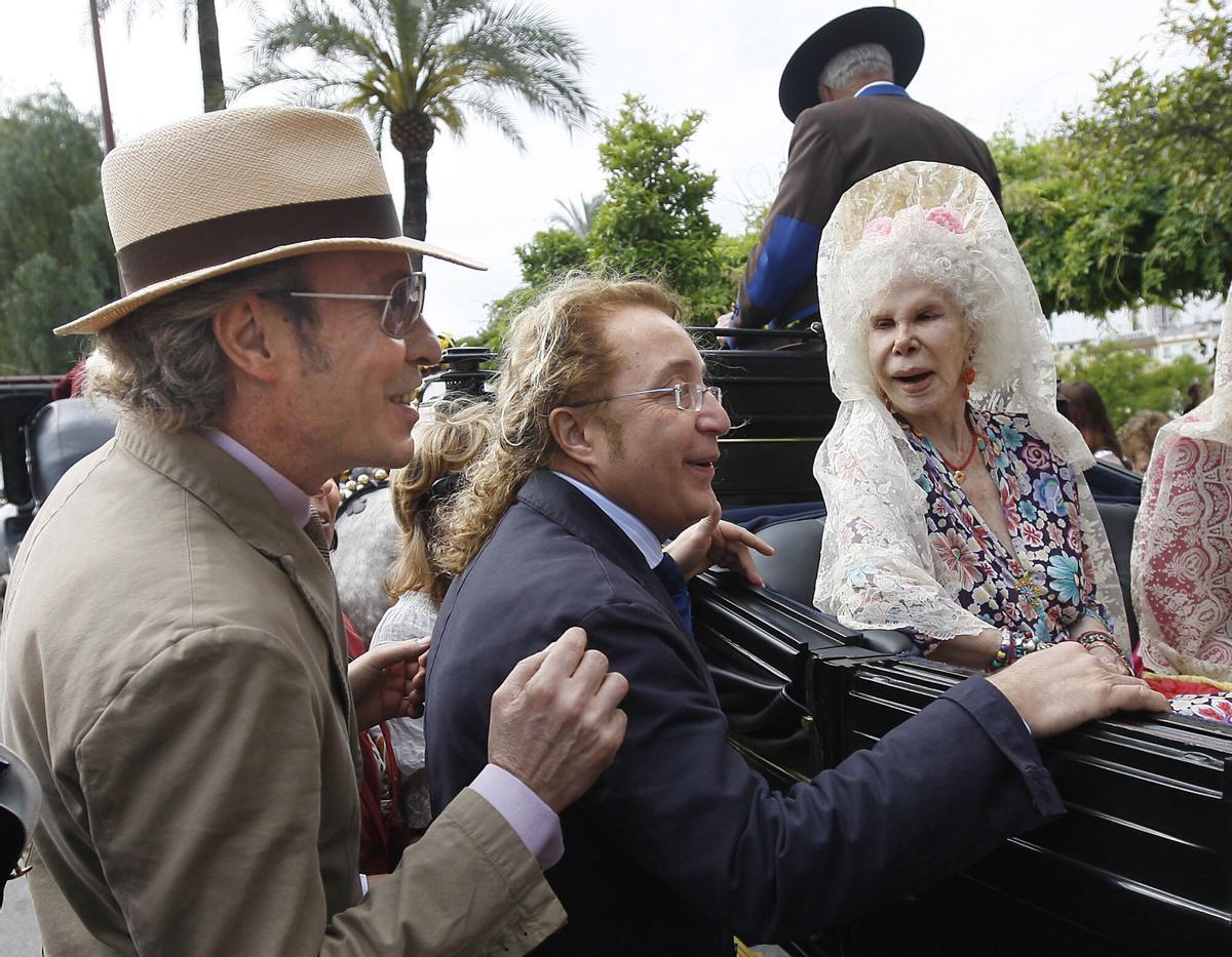 SEVILLA, 01/05/2011.- Los modistos José Víctor Rodríguez Caro (izda) y José Luis Medina del Corral (Vitorio y Luchino) saludan a la duquesa de Alba, Cayetana Fitz-James Stuart, en el carruaje en el que esta ha participado en la XXVI Exhibición de Enganches de la Feria de Sevilla, prevista en la plaza de toros de la Maestranza, pero que se ha tenido que celebrar en las calles cercanas al estar la arena de coso cubierta con una lona para evitar el deterioro del terreno ante la previsión de lluvias. EFE/Eduardo Abad ESPAÑA-FERIA SEVILLA