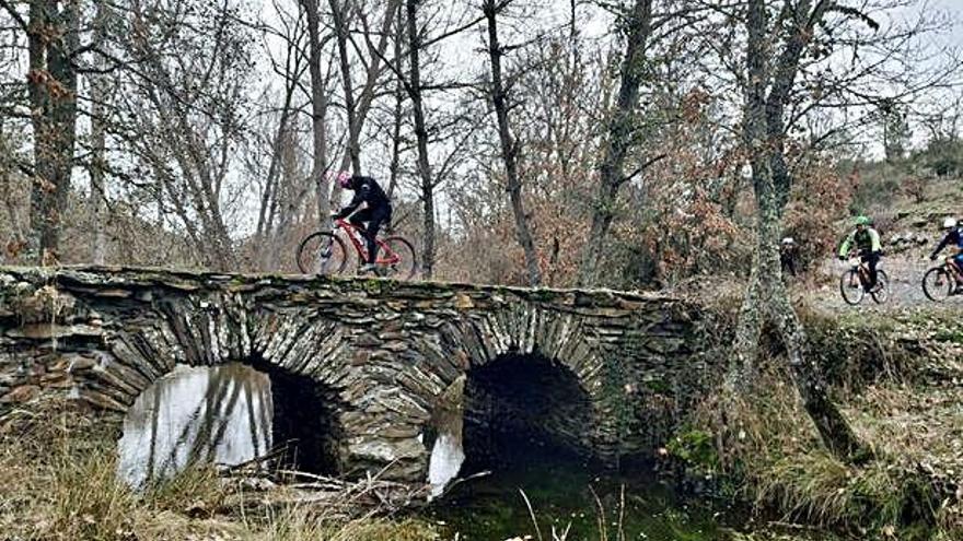 Un grupo de cicloturistas cruza un curso de agua por un antiguo puente de piedra.