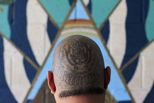 A leader of a powerful street gang with a tattoo on his head stands in front of a mural of El Salvador's coat of arms painted on the wall of a jail in Tonacatepeque