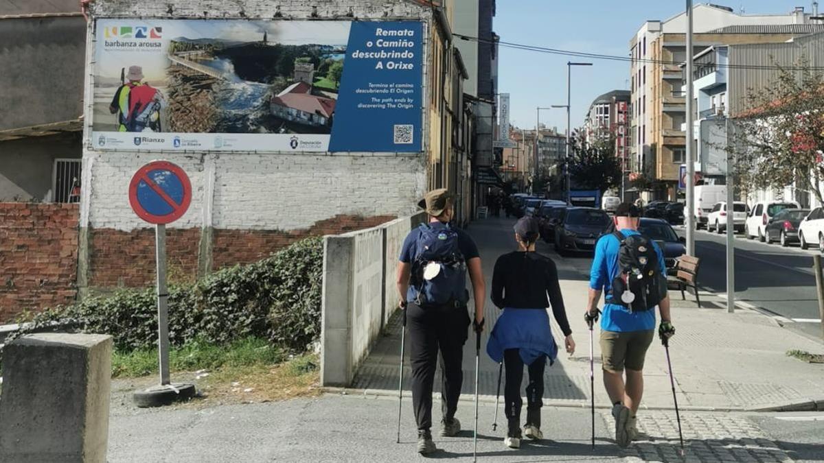 Tres peregrinos pasan en Arzúa ante el mural que promociona el Camino A Orixe.