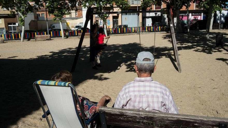 Dos jubilados, sentados en un parque mientras una niña juega en los columpios.