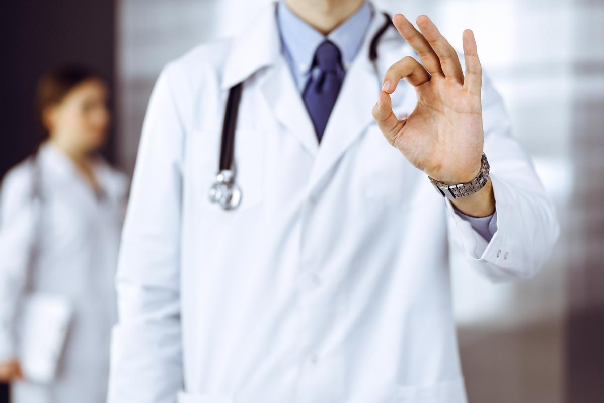 Unknown man-doctor standing straight and showing Ok sign in modern clinic. Female colleague is in a hurry at the background of physician. Medicine concept.