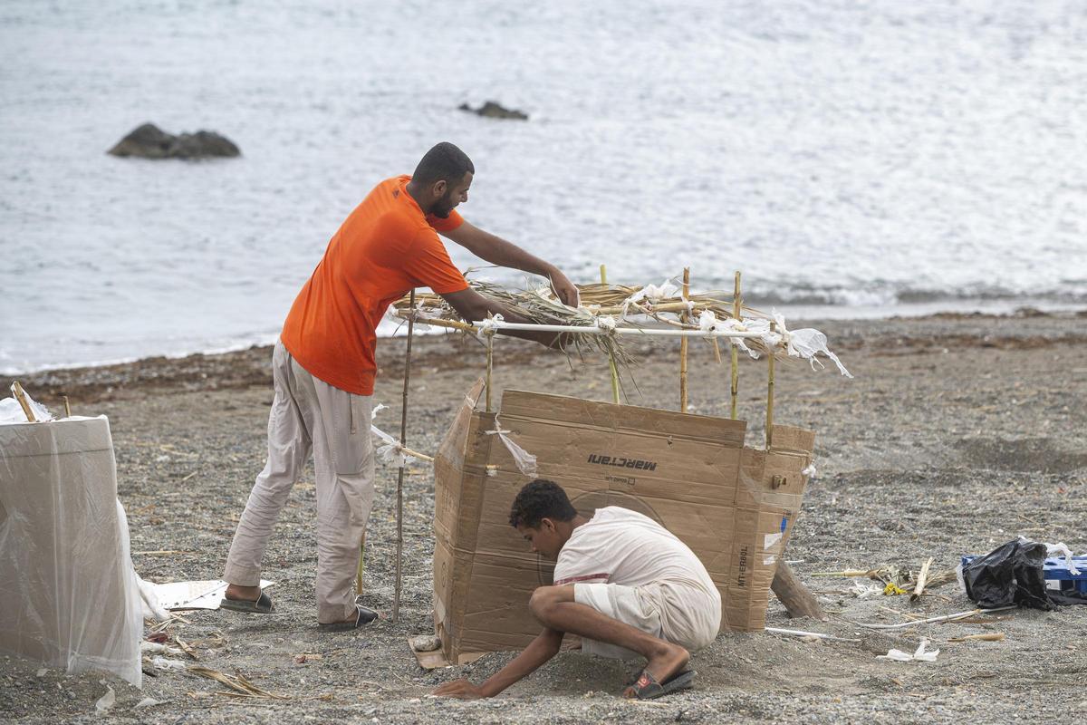 Varios migrantes vuelven a instalarse en la playa El Trampolín durante las tareas de limpieza, a 24 de agosto de 2026, en Ceuta (España). Operarios de limpieza se encargan de barrer y desinfectar el lugar tras el desalojo de la zona. Esta playa se ha conv
