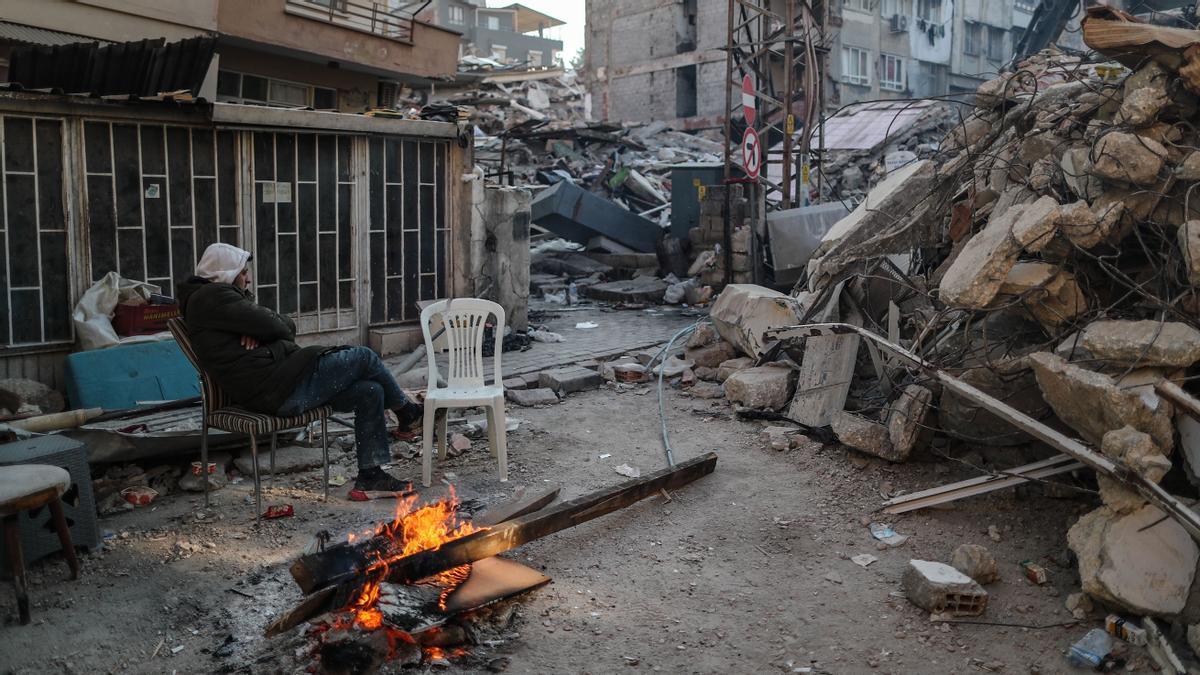 Un hombre sentado junto al fuego entre los edificios derrumbados tras el fuerte terremoto en Hatay 