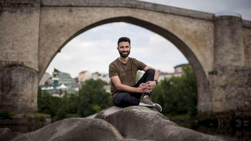 Cristian Fernández, sobre una roca en la ribera del Miño con el puente romano al fondo. // Brais Lorenzo