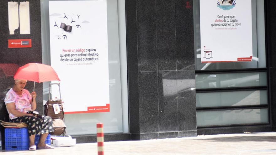 Una señora, sentada en la puerta de una sucursal bancaria de Murcia, se protege del sol con una sombrilla.