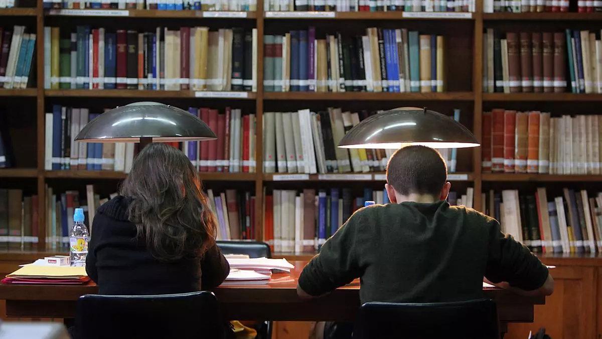 Dos estudiantes repasan sus apuntes en una biblioteca.