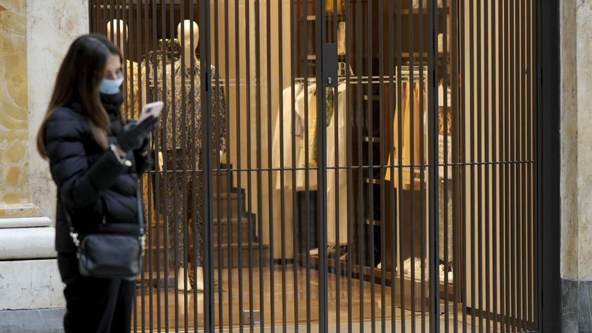 Una mujer con mascarilla manipula su teléfono móvil frente a una tienda de la Galería Umberto, en el centro de Nápoles, cerrada, el 11 de marzo del 2020