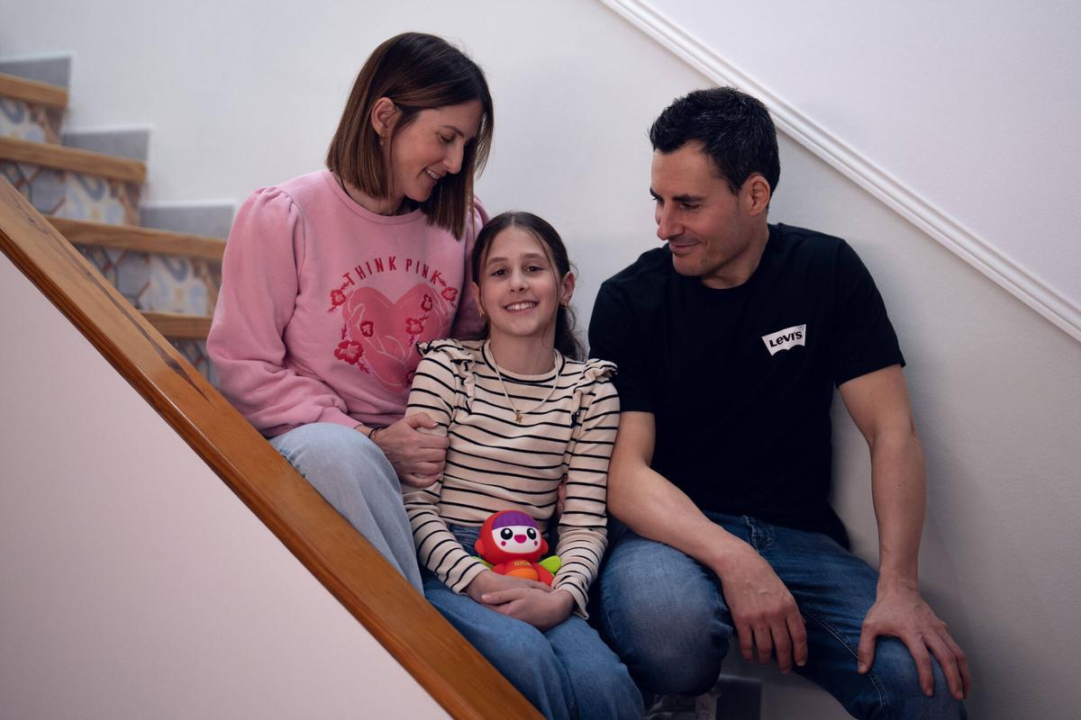 Martina, de 11 años y con una enfermedad rara, posa en la escalera de su casa junto a sus padres, Anna y Adrià, mientras sostiene una muñeca Nica.
