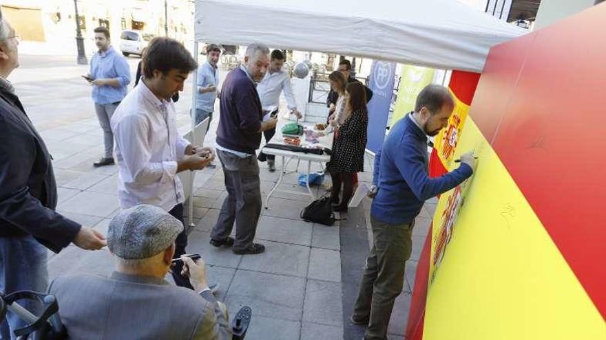 Arriba, un grupo de transeúntes firma en la bandera desplegada por el PP en Gijón. A la izquierda, Mercedes Fernández.