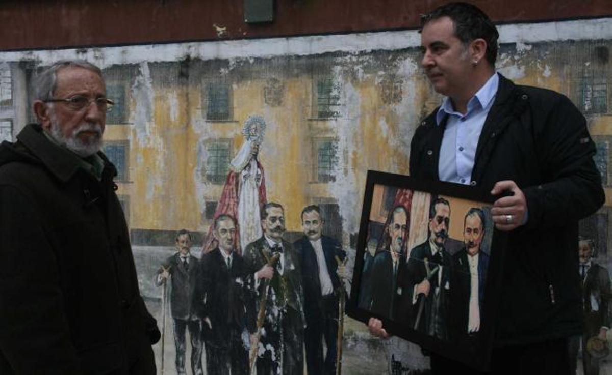 Alfredo Menéndez y Alberto López, que sostiene la muestra en foto cerámica de la misma escena que se representa a sus espaldas, ante el mural de Candás.