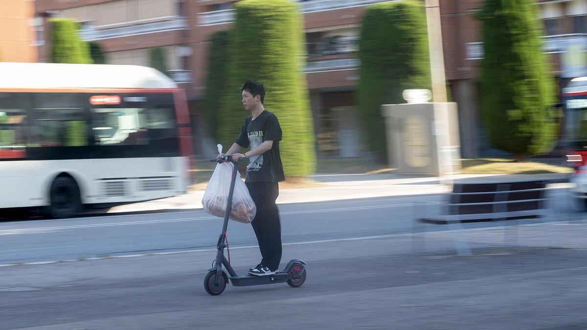 Un conductor de patinete circula cargado con bolsas por la plaza Mestres Maria Rosquellas i Joan Rotger de Badalona, junto a la estación de metro de Gorg antes de la nueva (y primera) regulación de circulación para patinetes eléctricos y demás VMP de la ciudad de Badalona