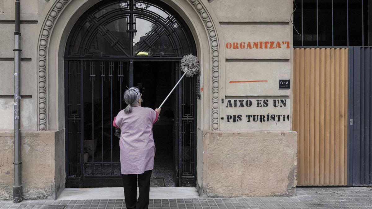 Una mujera limpia el portal en el que se lee una pintada que denuncia un piso turístico que hay en la finca en Barcelona.