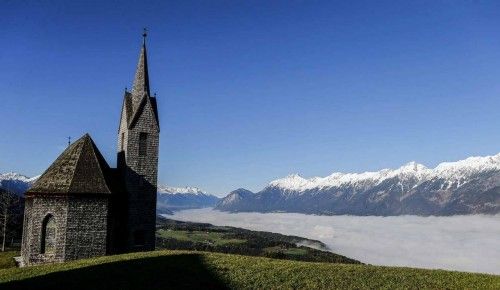 Una iglesia con vistas a un valle cubierto de niebla frente a unas montañas nevadas.