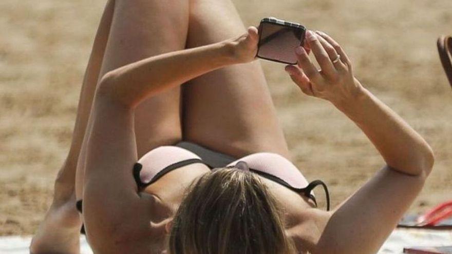 Una mujer mira su teléfono móvil mientras toma el sol en una playa.